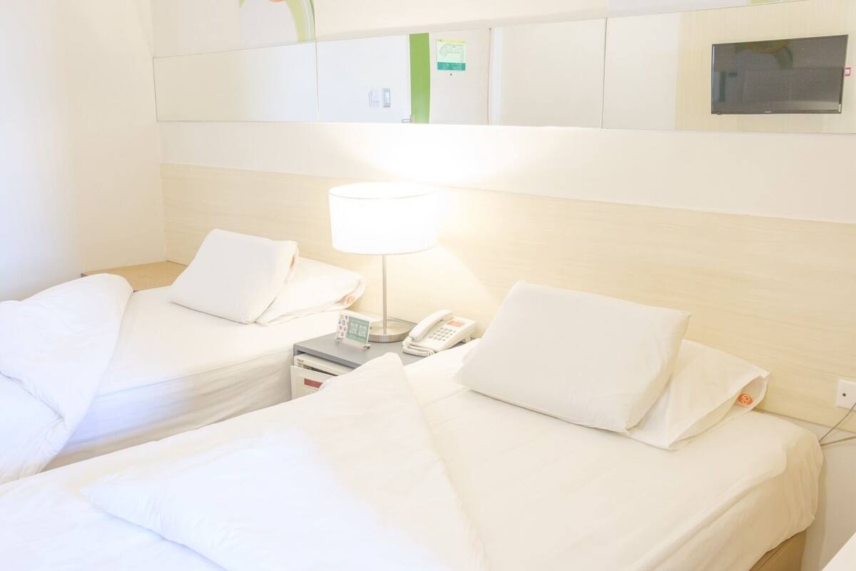 Ein Hotelzimmer mit zwei Einzelbetten, einem Nachttisch mit Lampe, einem Telefon und einem kleinen Schrank über den Betten.