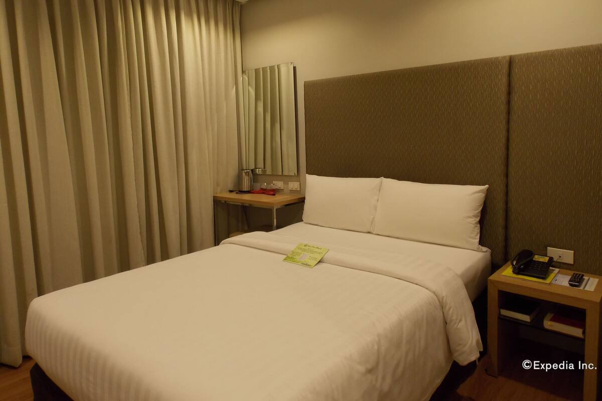 A hotel room with a large bed, a bedside table with a lamp, a mirror, and a small nightstand with a phone and books.
