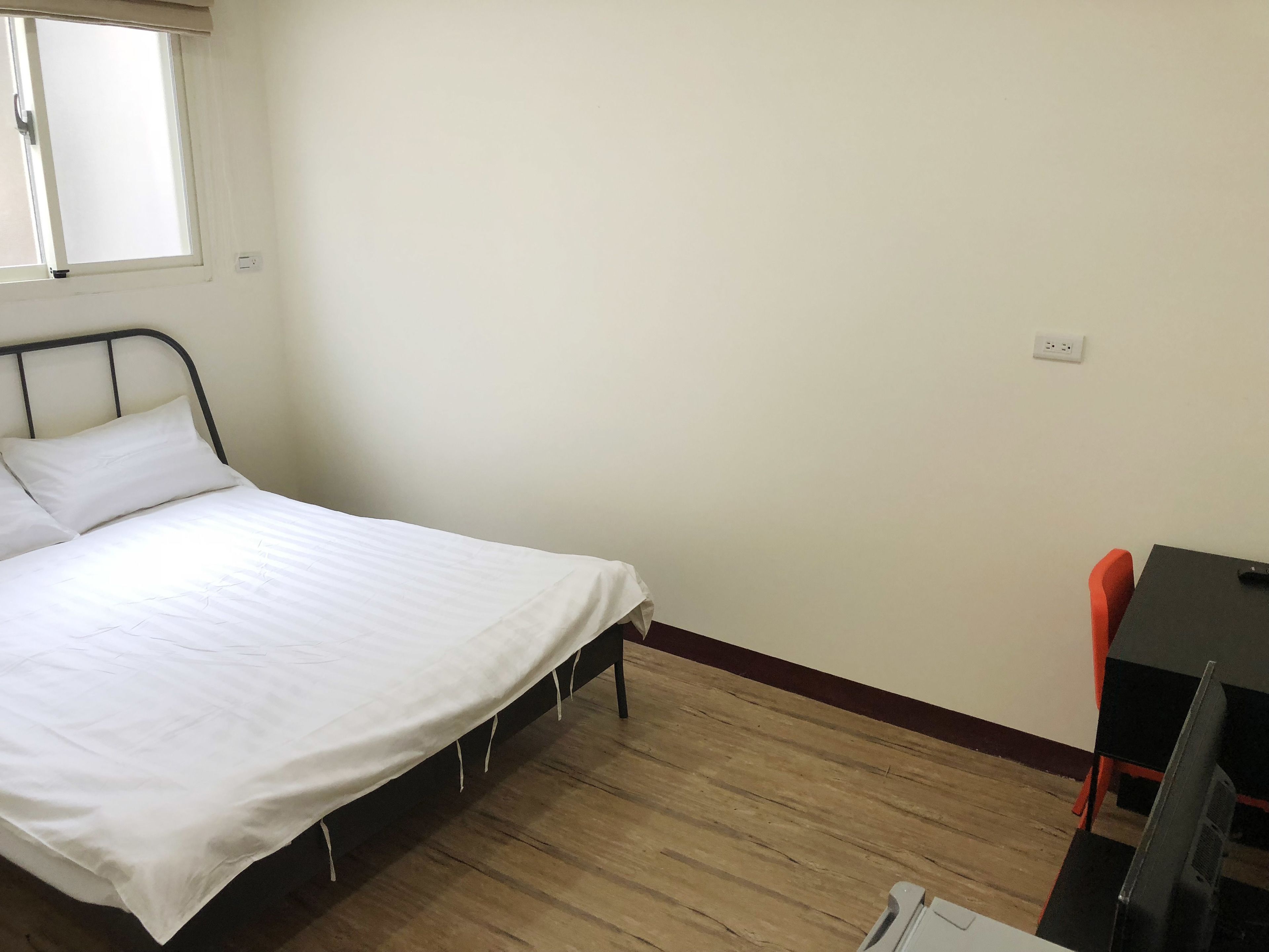 Una cama individual con ropa blanca en una habitación con suelo de madera, una mesita pequeña y una silla.