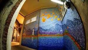 Ein schmaler Gang mit einer Mosaikwand, die eine Nachtszene mit Laternen zeigt.