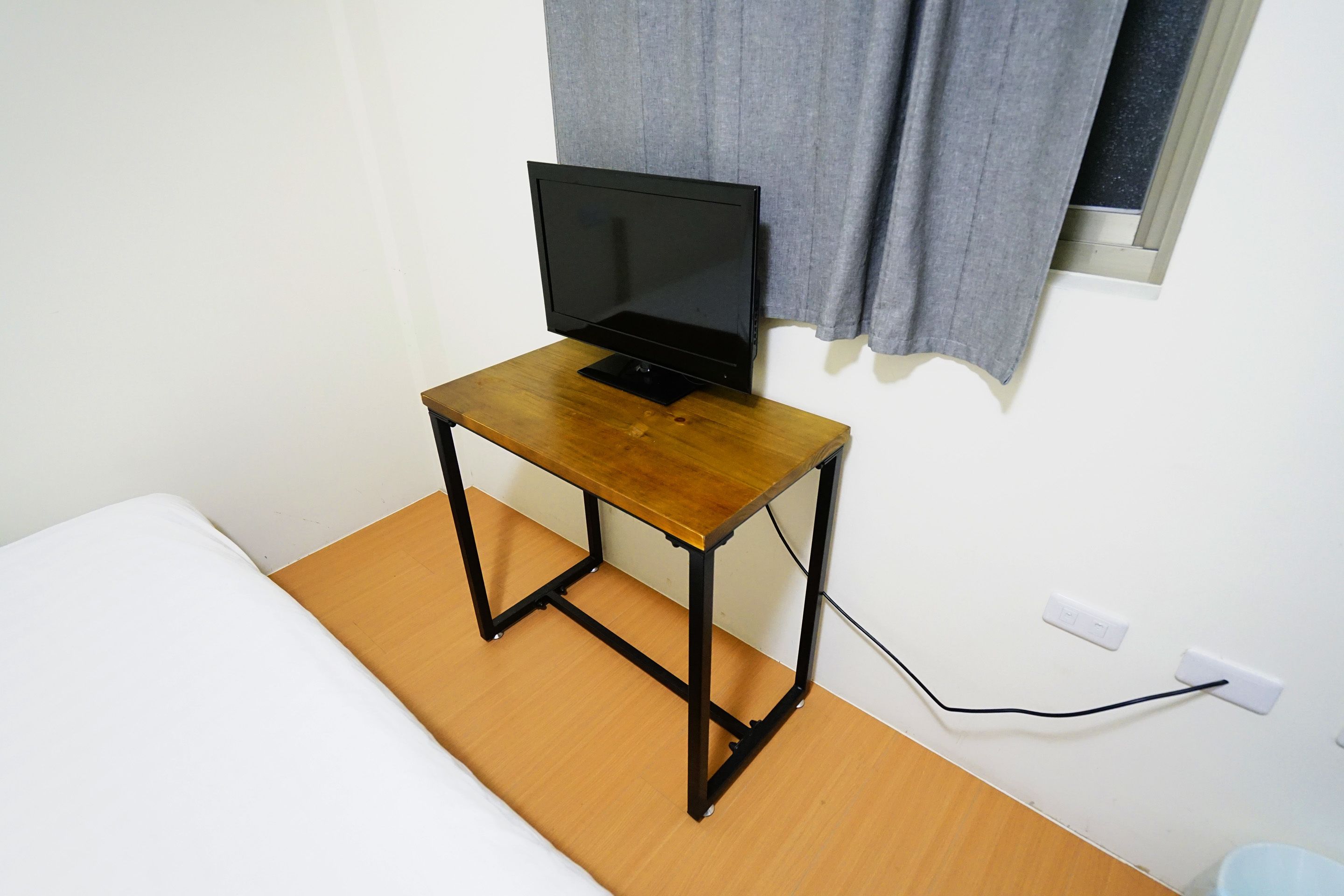 A small wooden table with a laptop on top, positioned next to a bed in a room with white walls and a window with curtains.