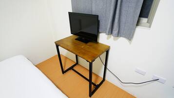 A small wooden table with a laptop on top, positioned next to a bed in a room with white walls and a window with curtains.