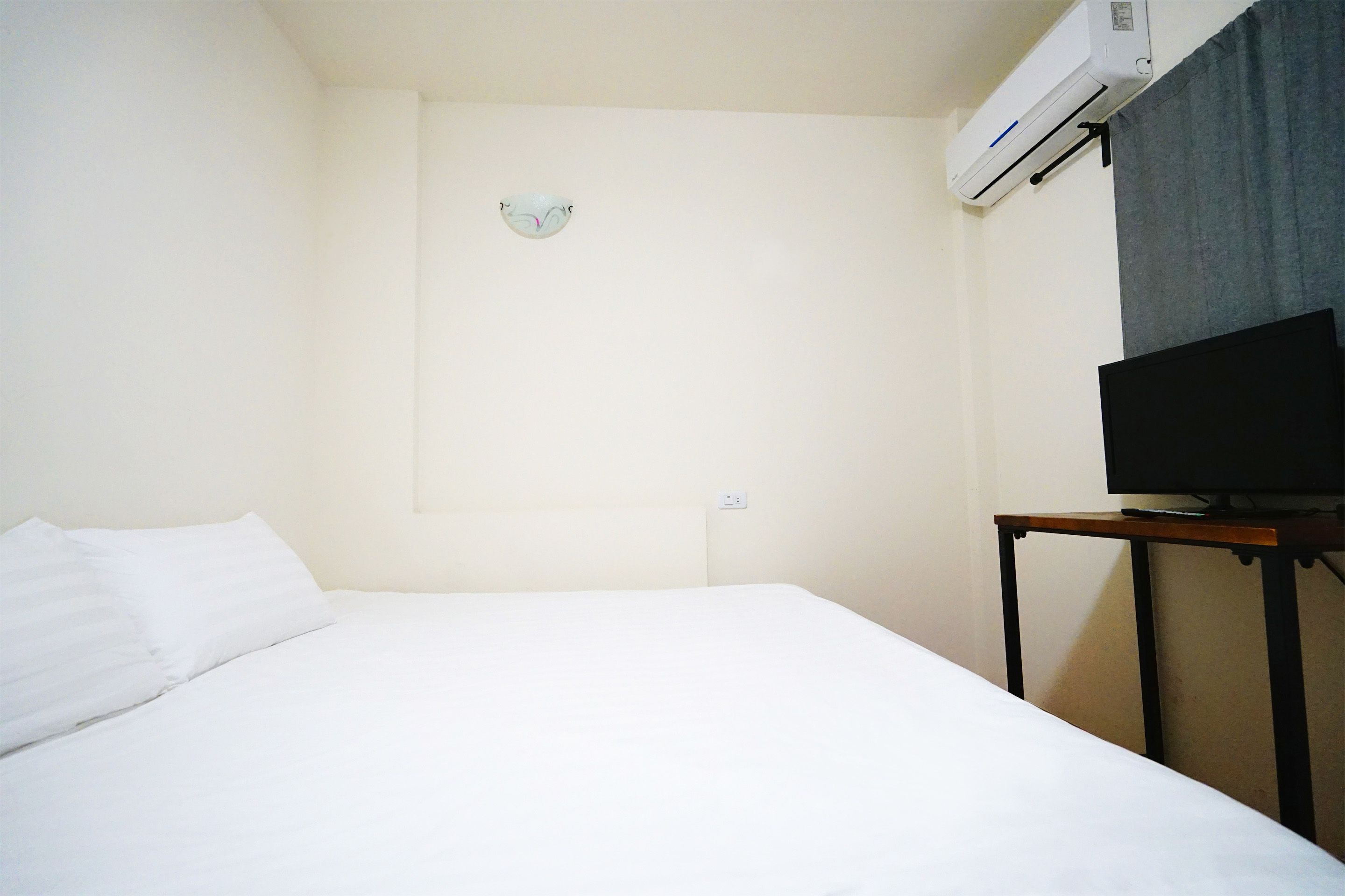 Quarto de hotel com cama, televisão sobre suporte de madeira e aparelho de ar condicionado fixado na parede.