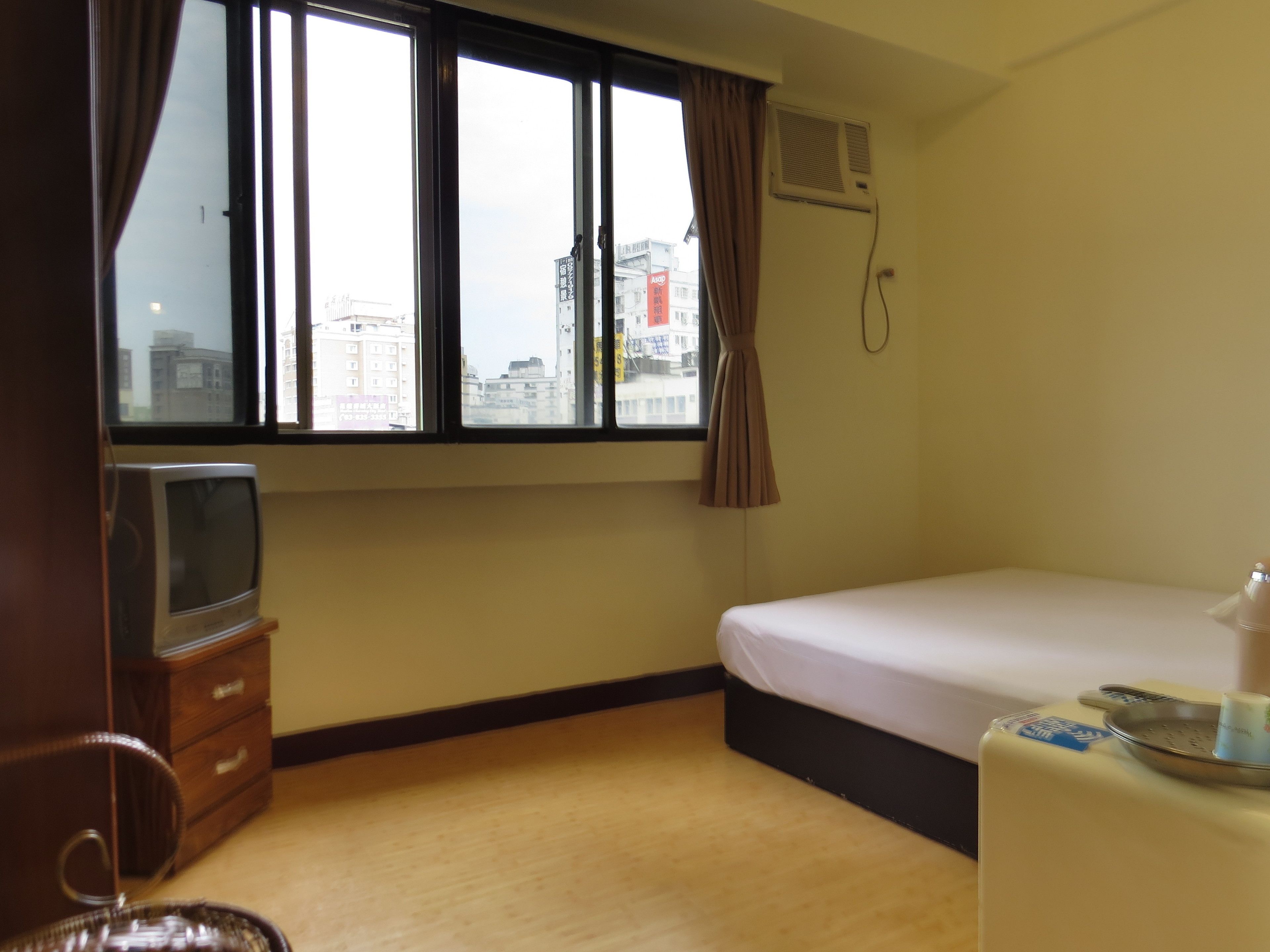A hotel room with a bed, a TV on a wooden cabinet, a window with curtains, and a view of buildings outside.