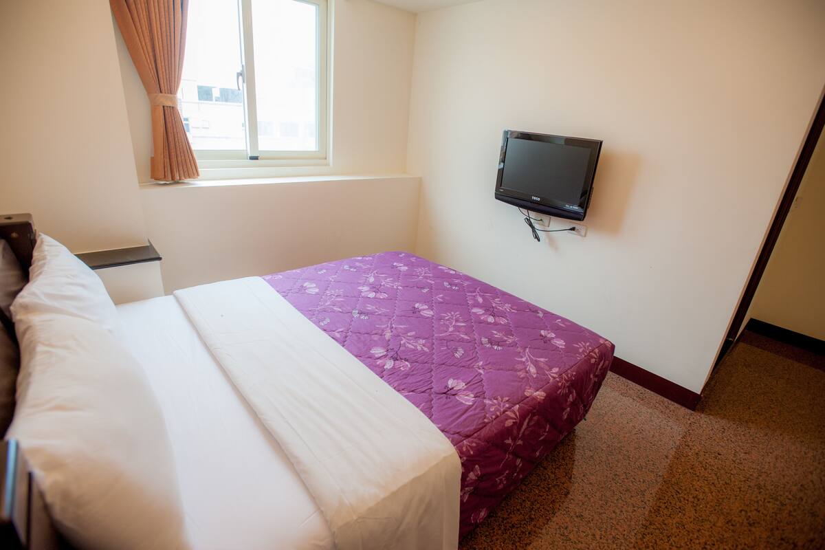 A bedroom with a bed, a TV mounted on the wall, and a window with curtains.
