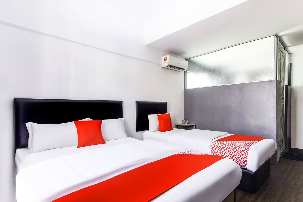 A hotel room with two beds, each with a black headboard, white bedding, and red accents. There is a small table and a chair, and an air conditioning unit mounted on the wall.