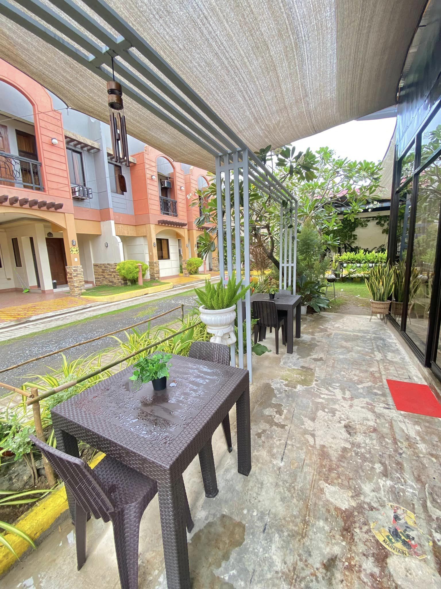Un espacio al aire libre techado con una mesa y sillas, plantas en macetas y vistas a la calle y edificios al fondo.