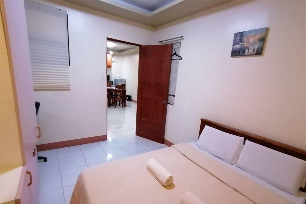 Um quarto com uma cama, uma porta de madeira, um quadro na parede e um corredor que dá para uma sala de jantar.