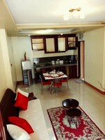 Una sala de estar con un sofá, cojines rojos, una mesa de centro y una alfombra. Se observa un comedor con una mesa dispuesta para cuatro personas y sillas rojas. En el fondo, se distingue una cocina con armarios y un microondas.