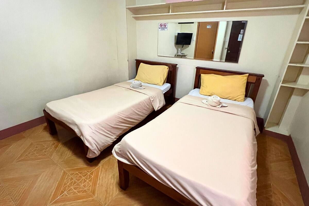 Duas camas de solteiro com estrutura de madeira e roupa de cama bege, um espelho acima e um aviso de 'Não Fumar' na parede.
