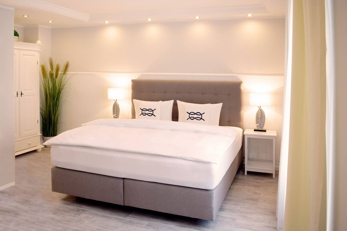 Een moderne slaapkamer met een groot bed, twee bedlampen, een nachtkastje en een hoge plant.