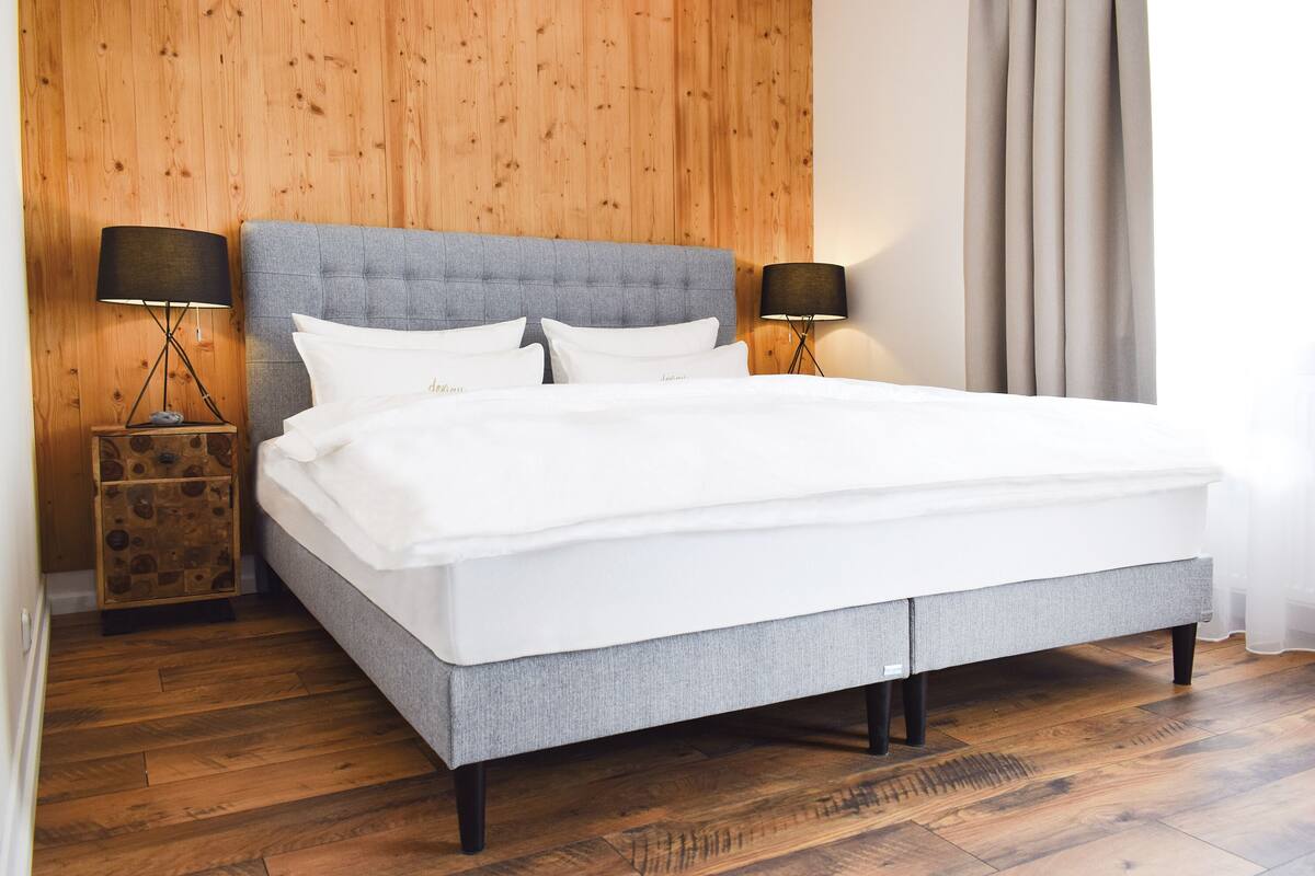 Een moderne slaapkamer met een grijs gestoffeerd bed, twee nachtkastjes met lampen, houten vloeren en een houten wandpaneel.