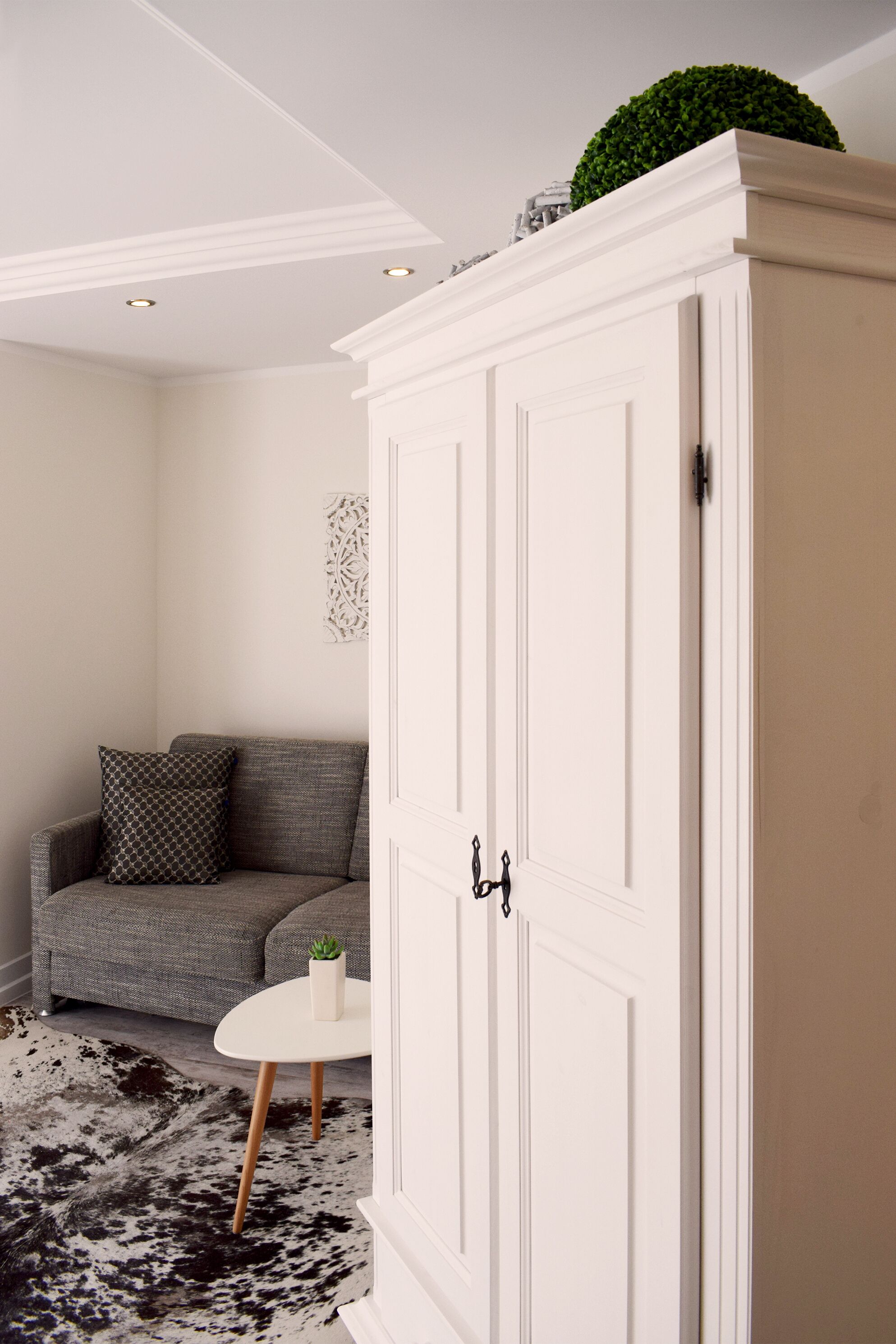 Une pièce avec une armoire blanche, un canapé gris, une petite table blanche et un tapis en peau de vache.