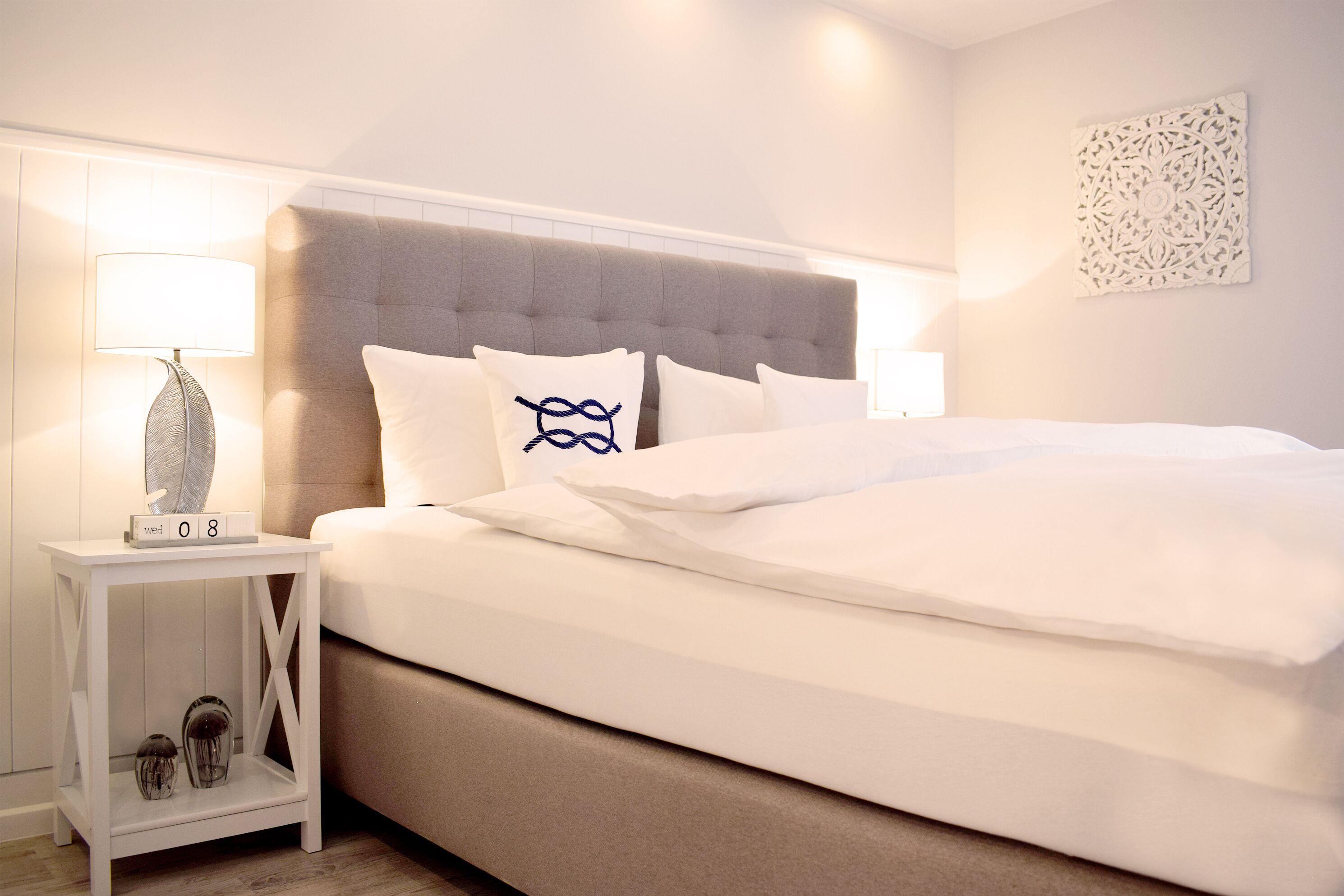 Una cama bien hecha con ropa de cama blanca y un cabecero gris, una mesita de noche con una lámpara y un adorno en la pared.