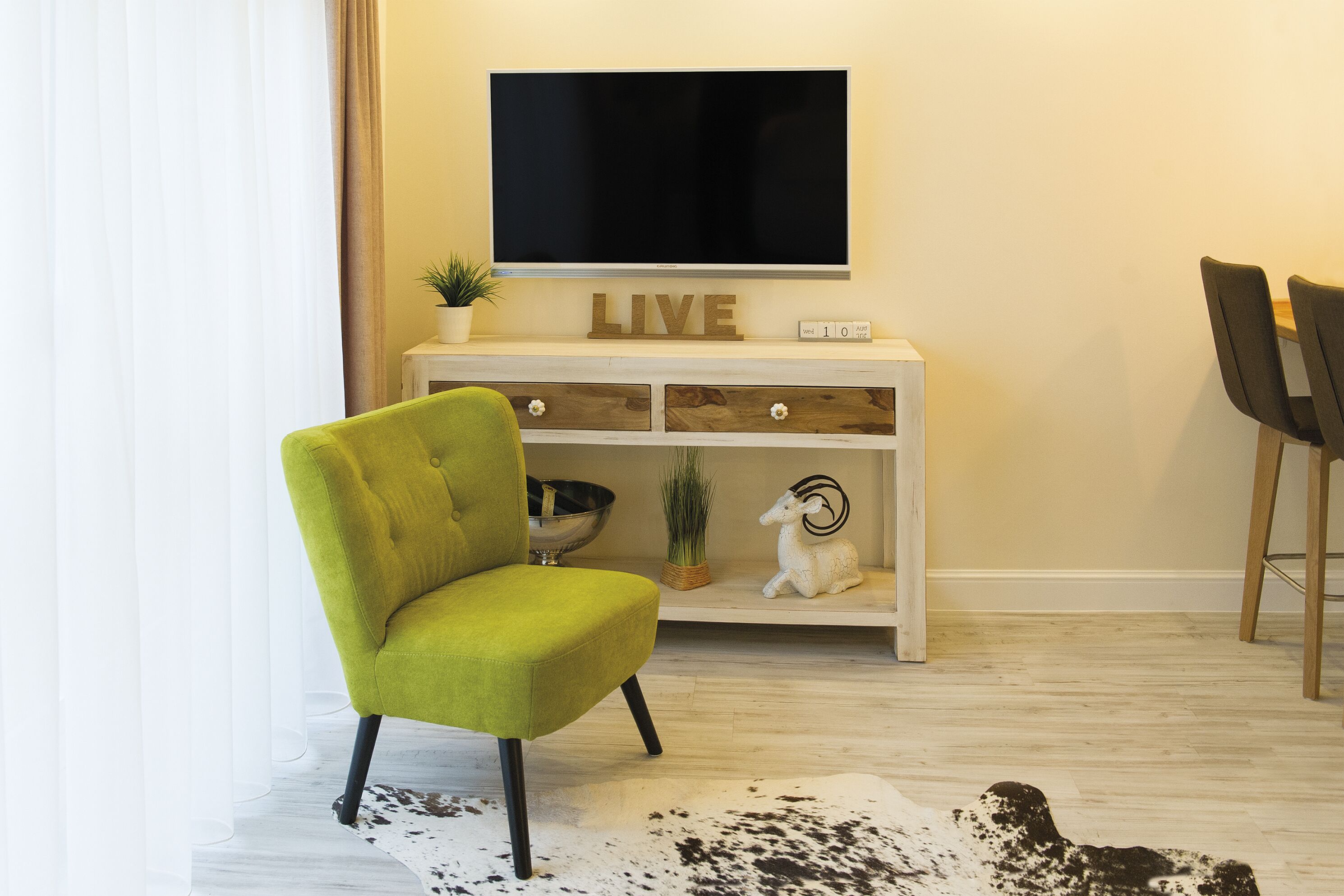 Un salon avec un fauteuil vert, un téléviseur à écran plat fixé au mur, une console en bois ornée d’objets décoratifs et un tapis en peau de vache sur le sol.