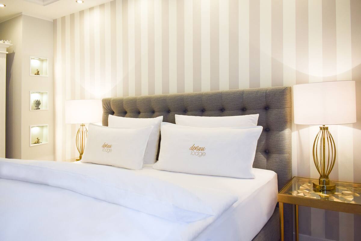 Een netjes opgemaakt bed met witte lakens en kussens, een gestofte hoofdbord en twee bedlampjes op een gouden nachtkastje.