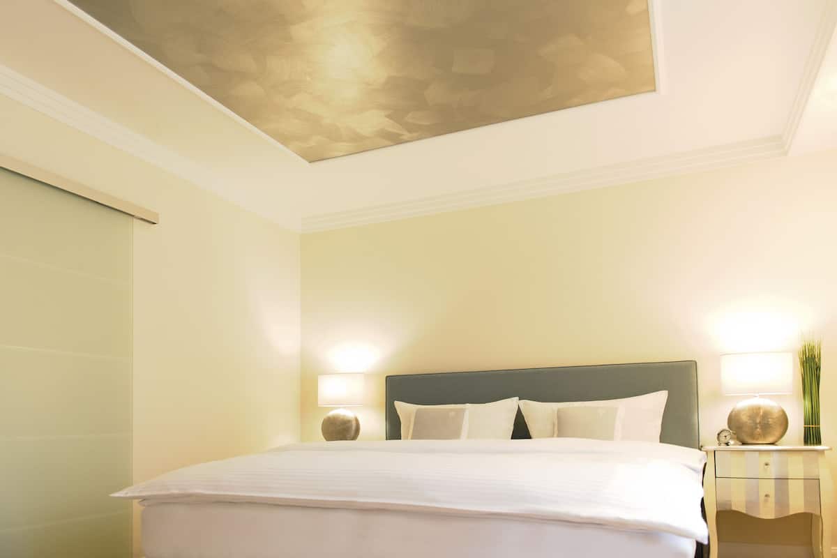Een net opgemaakt bed in een hotelkamer, met een modern hoofdbord, nachtlampjes en een decoratief wandobject.