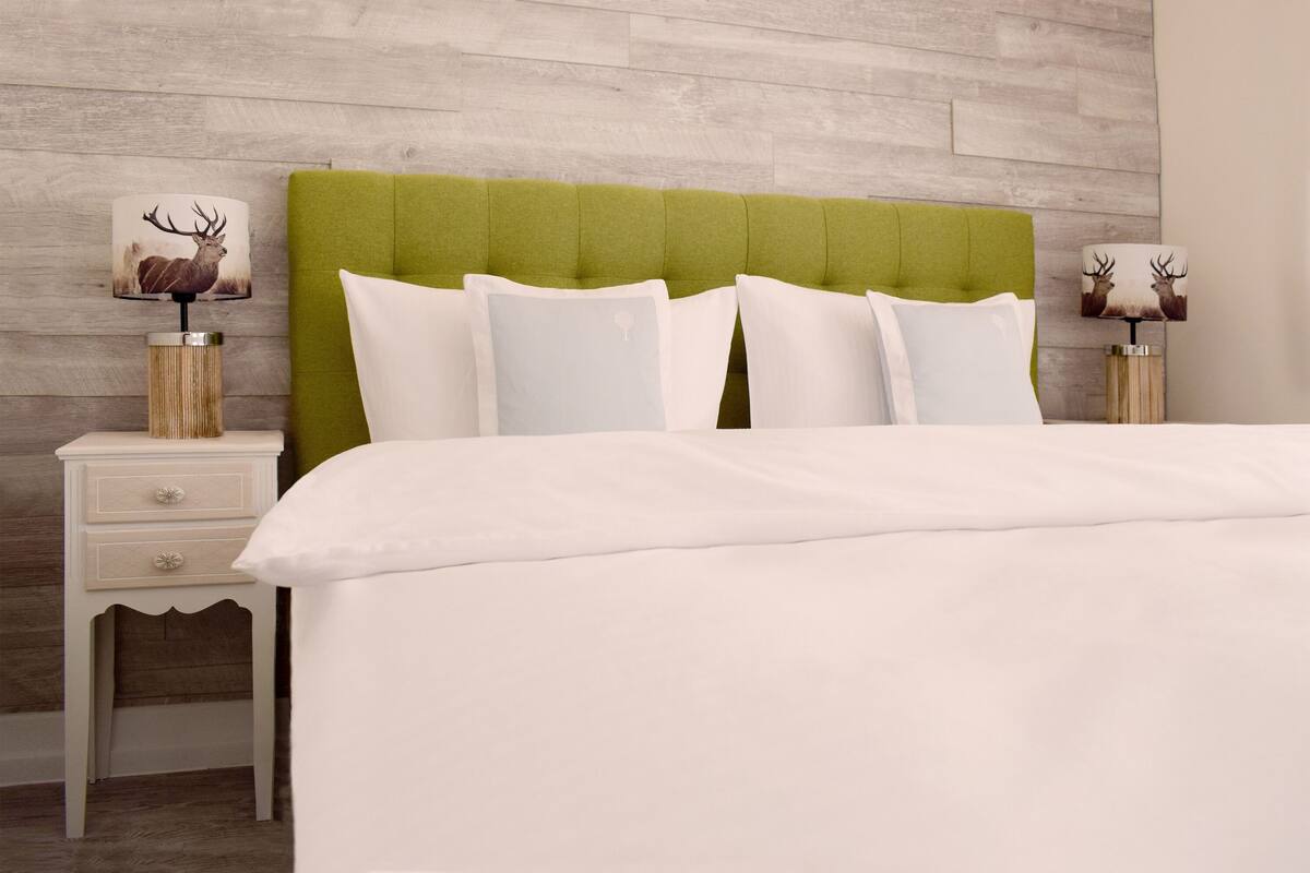 Een net opgemaakt bed met een groen beklede hoofdeinde, witte beddengoed en decoratieve lampen met hertmotief aan weerszijden.
