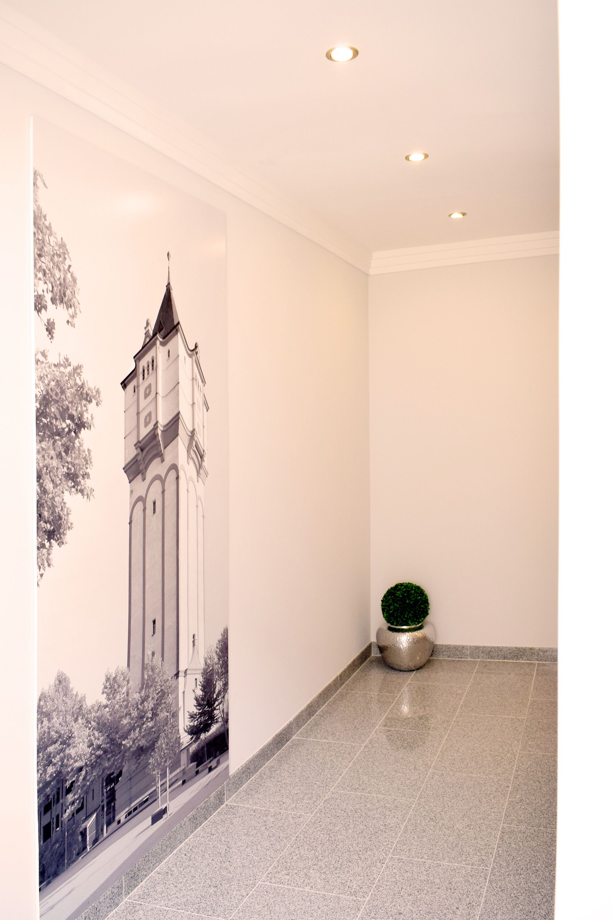 Un couloir avec un sol carrelé, un tableau représentant un bâtiment accroché au mur et une petite plante en pot.