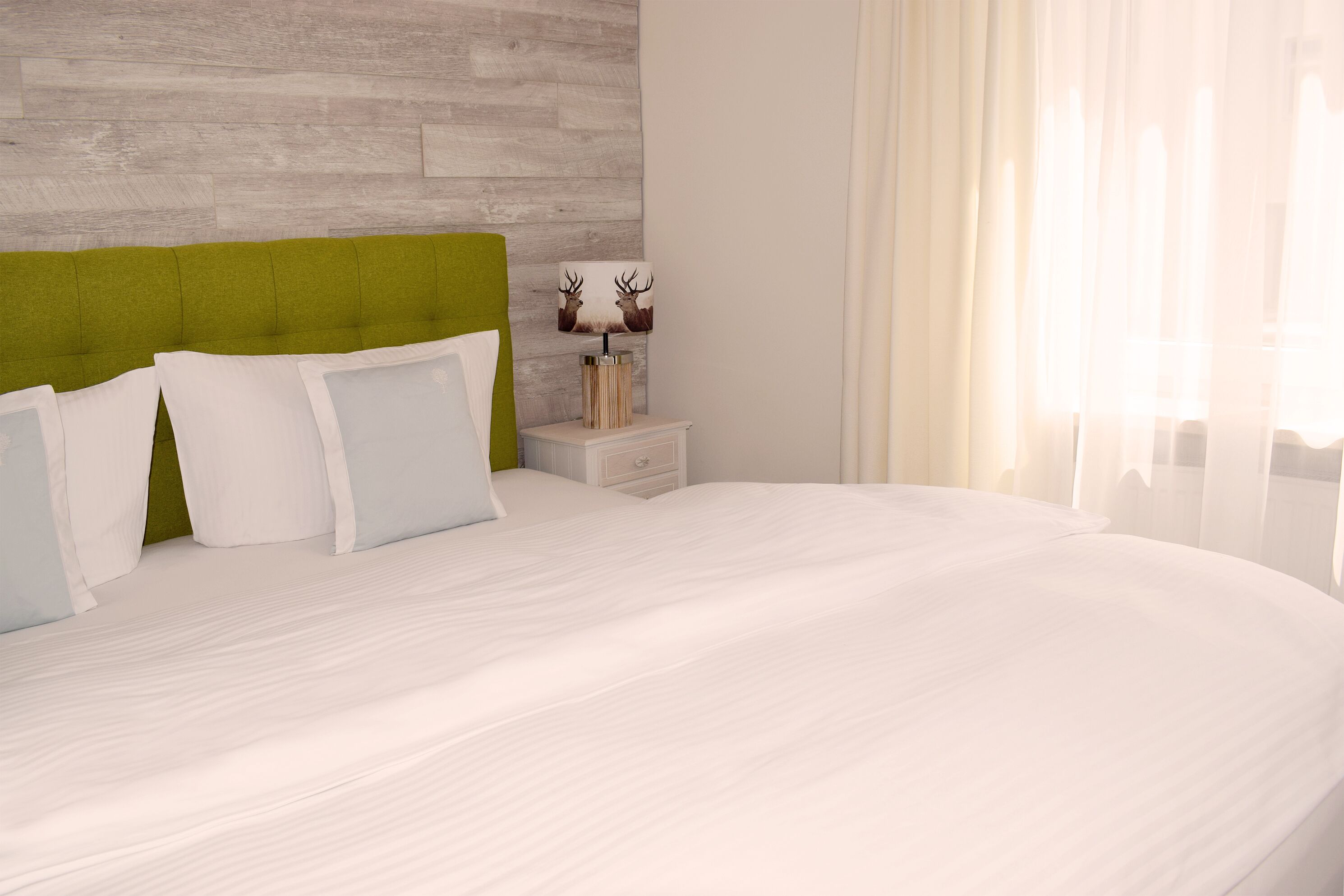 Una cama bien tendida con sábanas blancas, un cabecero tapizado en verde y una mesita de noche con una lámpara y objetos decorativos.