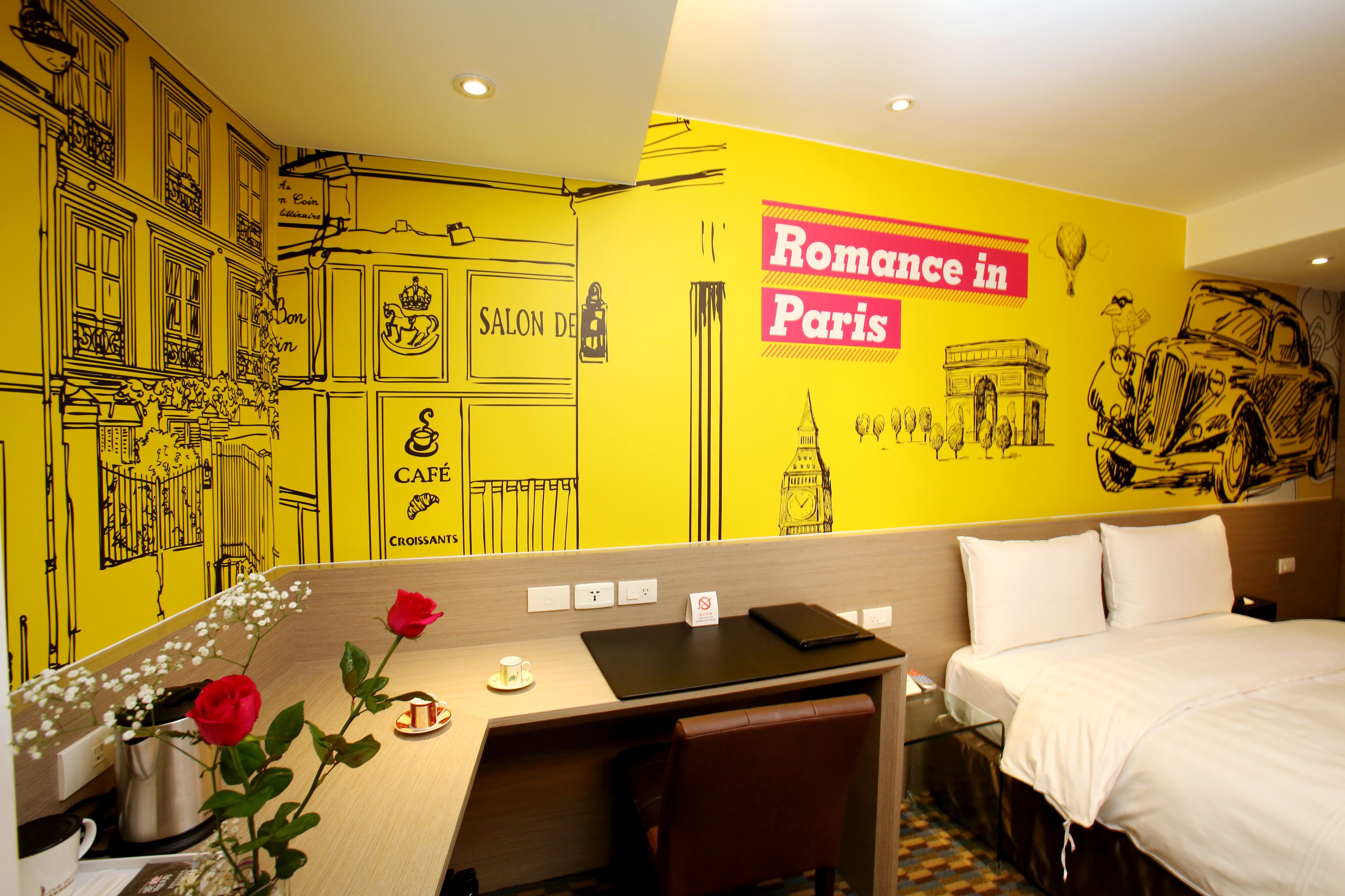 A hotel room with a vibrant yellow wall, a bed, a desk with a laptop, and a small table with a vase of flowers.