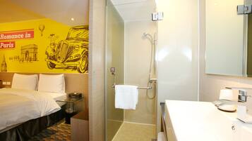 A hotel room with a bed, a glass-enclosed shower, a sink, and a mirror.