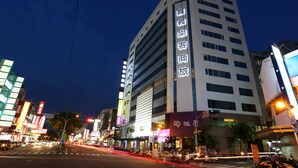 Un hotel de varios pisos con señalización iluminada, un paso de peatones y edificios urbanos circundantes.