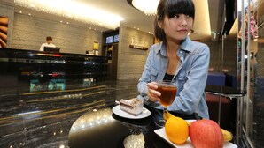 Una mujer sentada a una mesa con una porción de pastel, una taza de té y fruta. El entorno parece ser el vestíbulo de un hotel con un mostrador de recepción y una araña.