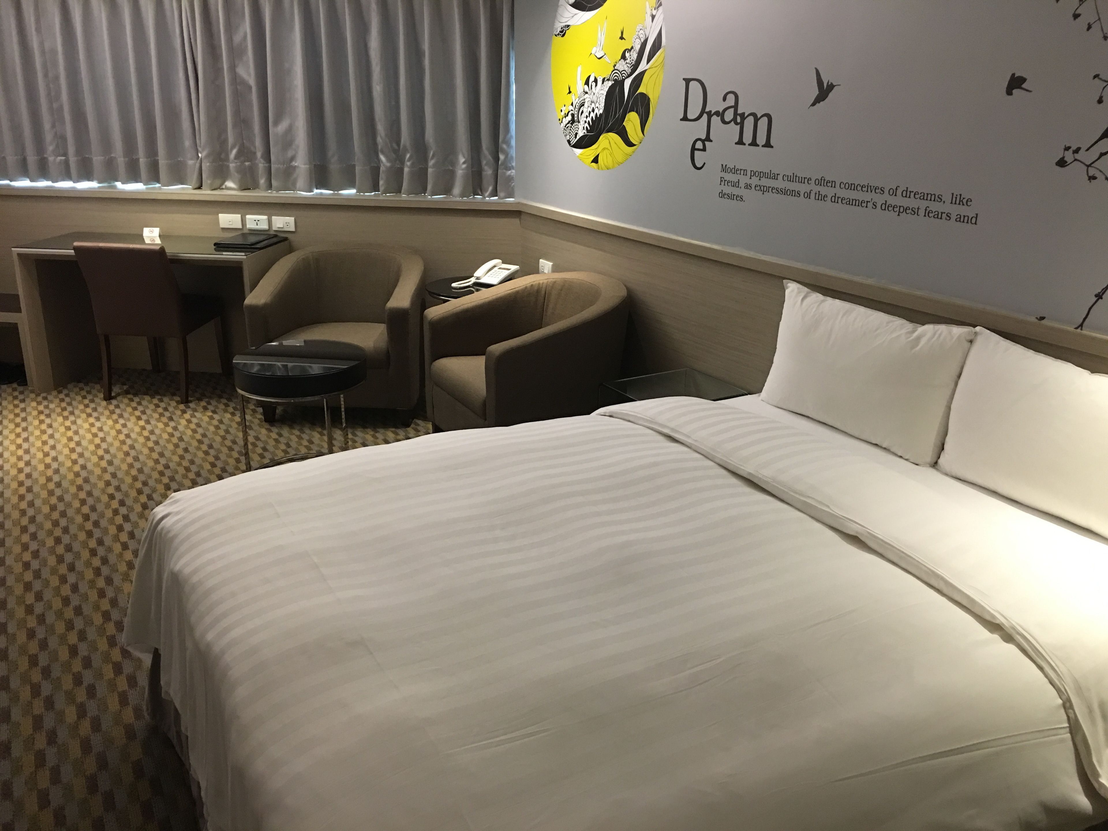 A hotel room with a large bed, a desk, two chairs, a small table, and a wall with a poster.