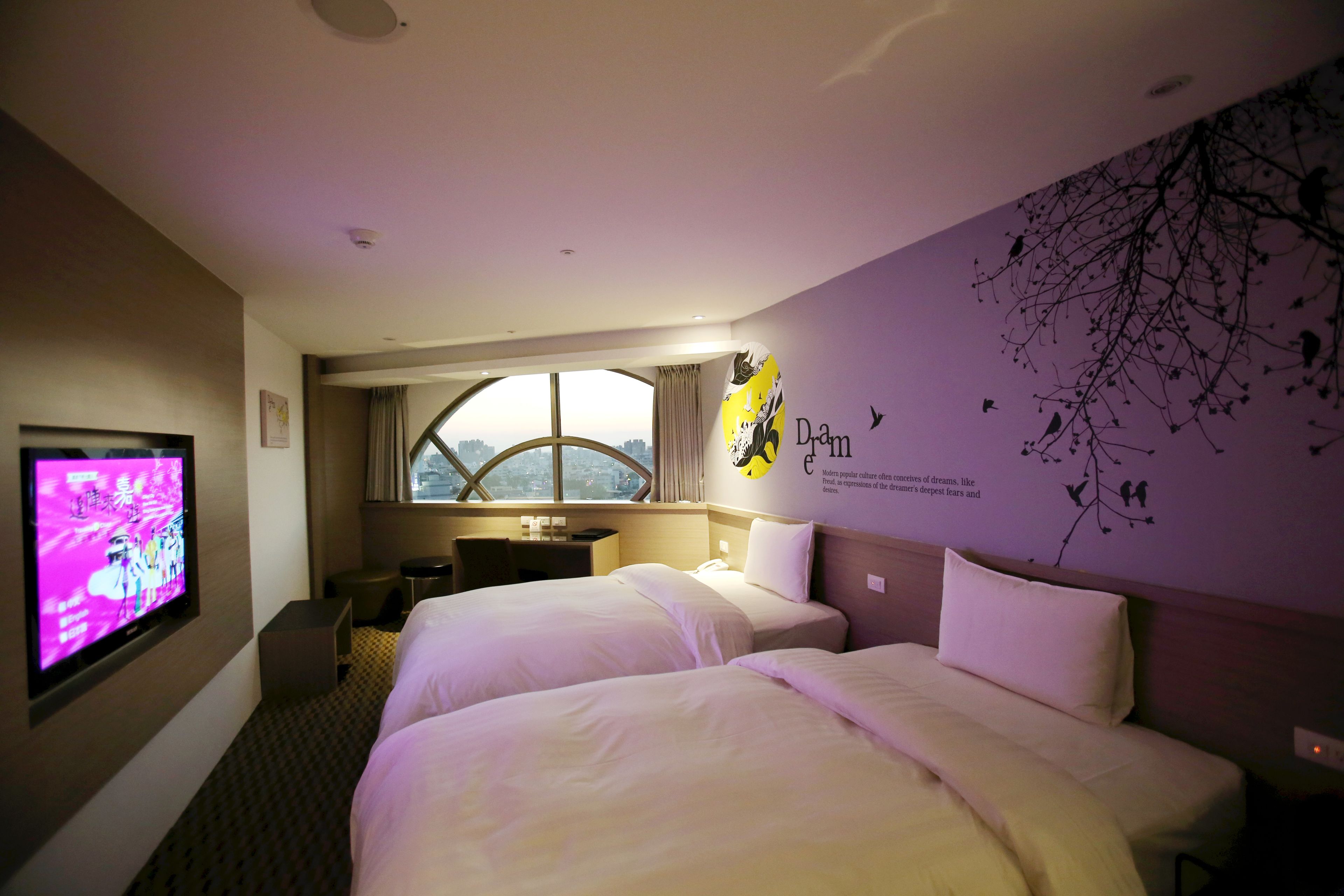 A hotel room with two beds, a TV, a desk, and a window with a view.