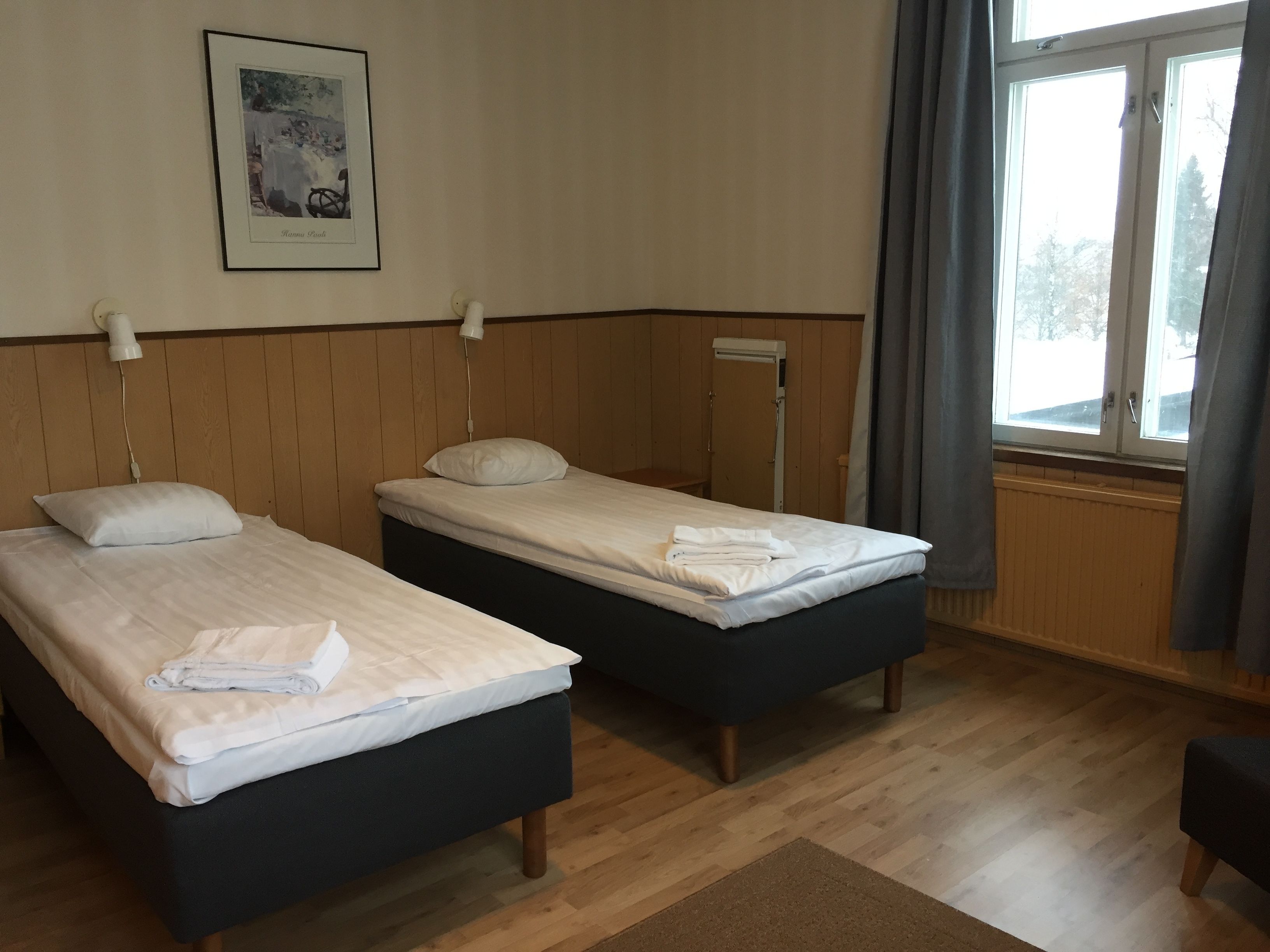 Quarto de hotel com duas camas de solteiro, piso de madeira, janela com cortinas e um quadro na parede.