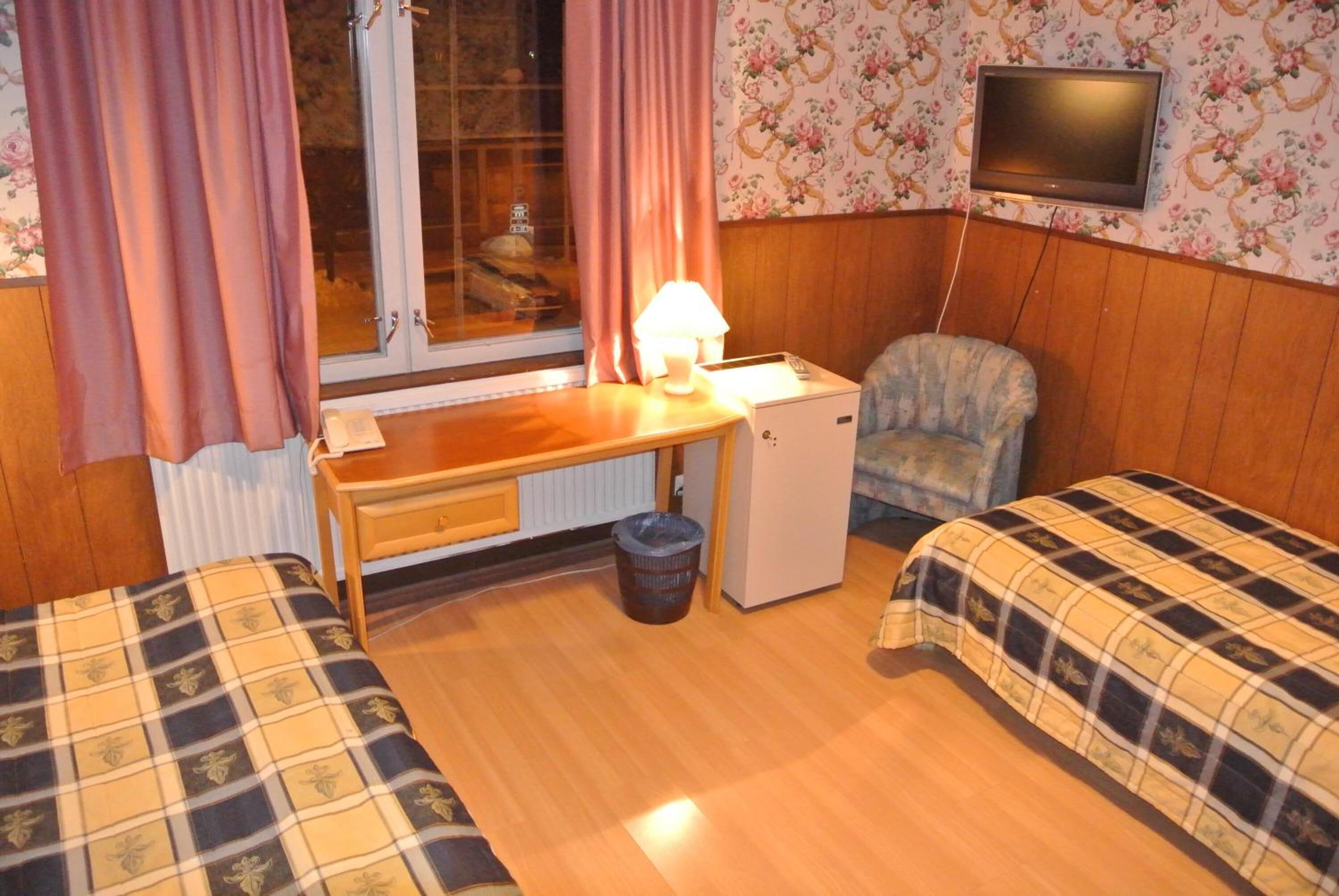 A small room with two beds, a desk, a chair, a television, and a window with curtains.
