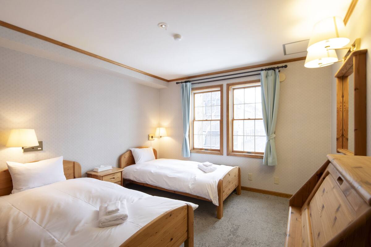 Una habitación de hotel con dos camas individuales, un aparador de madera, una mesita de noche con lámpara y una ventana con cortinas.