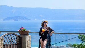 A person in a black swimsuit and hat, holding a drink, standing by a railing with a view of the sea and mountains. - Sunny Villas and Spa (Kassandra)
