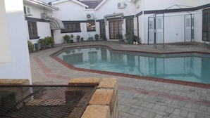 A swimming pool surrounded by a courtyard with white buildings, potted plants, and a metal fence. - White Palace Hotel (Serowe)