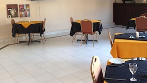 A dining area with tables set for two, a flat-screen TV mounted on the wall, and a kitchen area in the background. - White Palace Hotel (Serowe)
