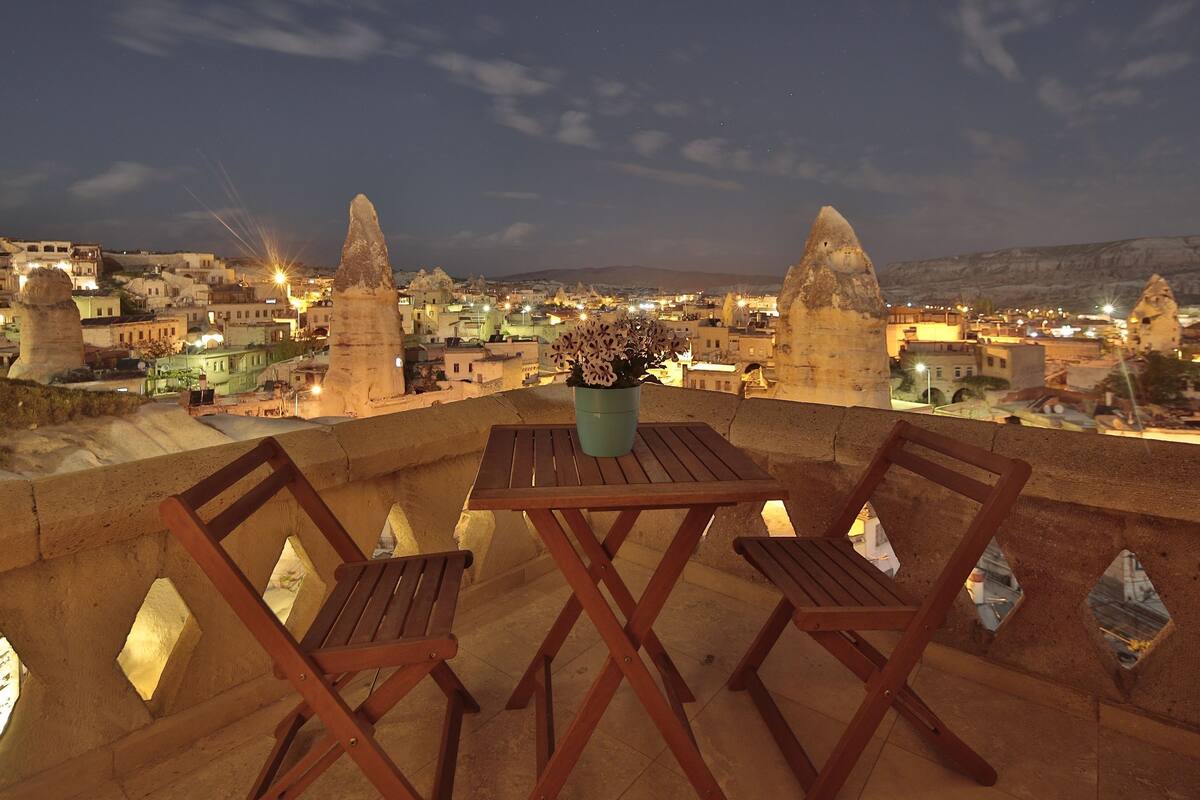 A terrace with two wooden chairs and a table, overlooking a town with distinctive rock formations and illuminated buildings.