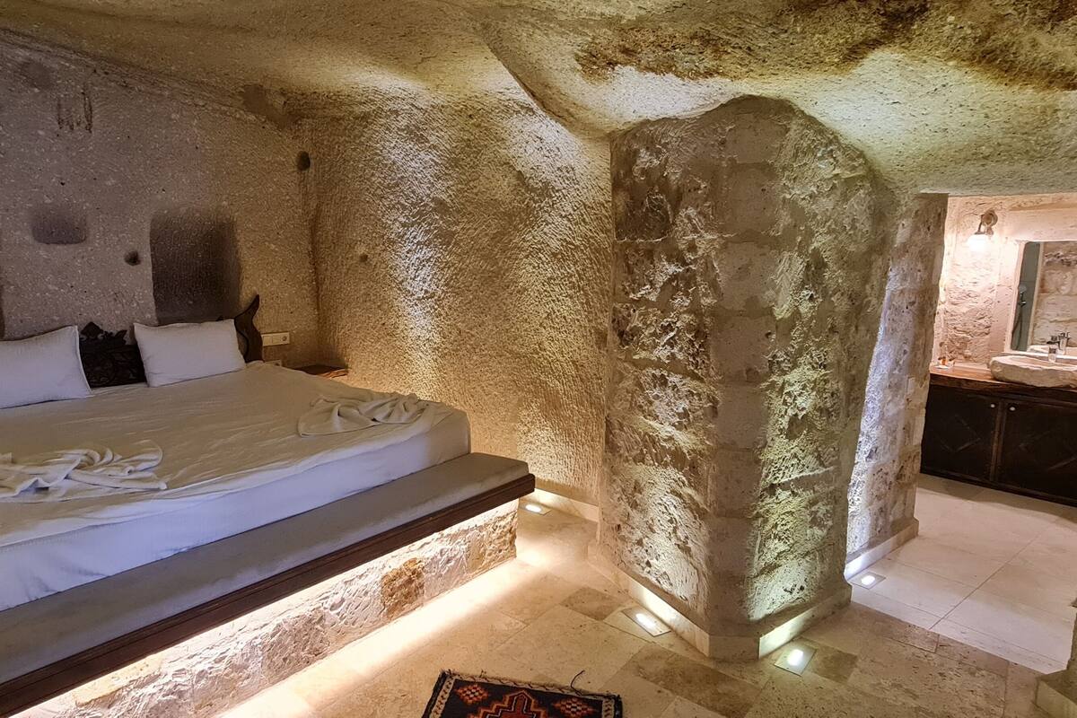 A bedroom with a stone wall, a bed, a nightstand, and a bathroom with a mirror and sink.
