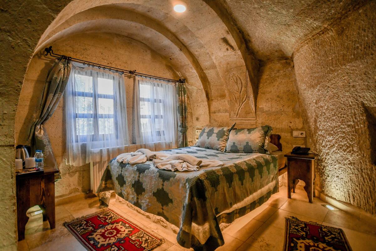 A bedroom with a bed, a nightstand, a window with curtains, and a stone wall.