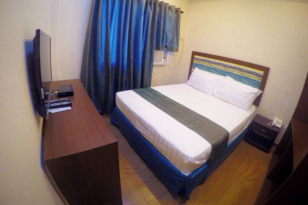 Habitación de hotel con cama, escritorio, computadora portátil, teléfono y ventana con cortinas.