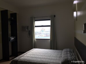 A hotel room with a bed, a window with curtains, a wall-mounted air conditioning unit, and a view of a building outside.