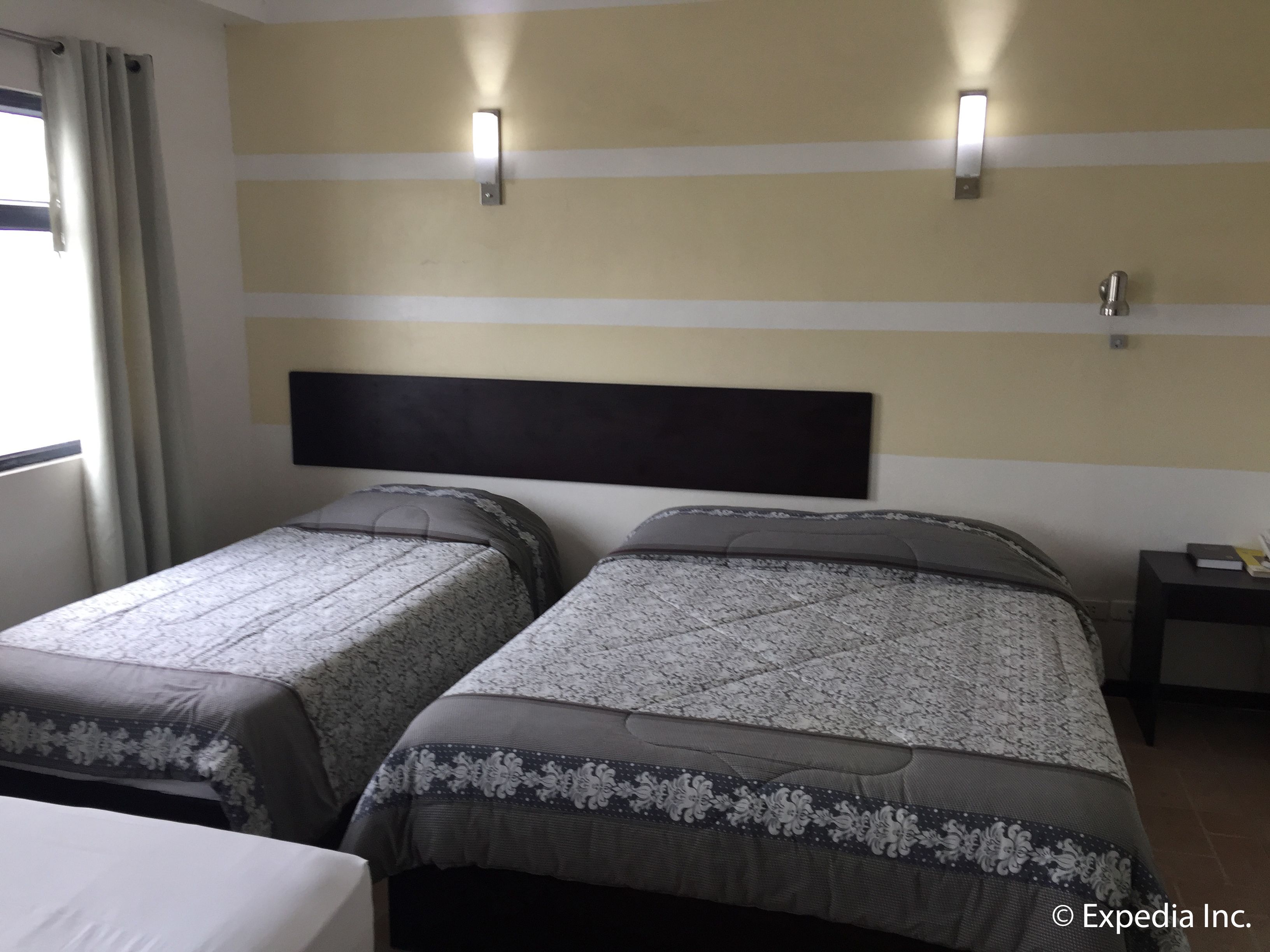 A hotel room with two beds, each with a headboard and a decorative pillow. There are two wall-mounted lights and a small table with a book.