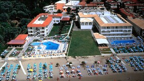 An aerial view of a coastal resort with multiple buildings, a central swimming pool, and numerous beach umbrellas and sun loungers. - Astir Beach (Zakynthos)