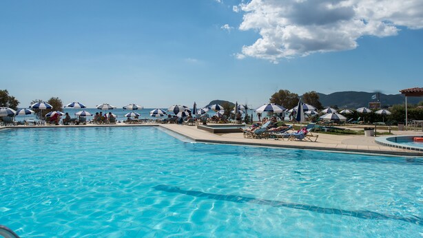 A pool area with multiple lounge chairs and umbrellas, a clear sky, and a view of the sea and mountains in the background. - Astir Beach (Zakynthos)