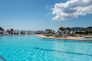 A pool area with multiple lounge chairs and umbrellas, a clear sky, and a view of the sea and mountains in the background.