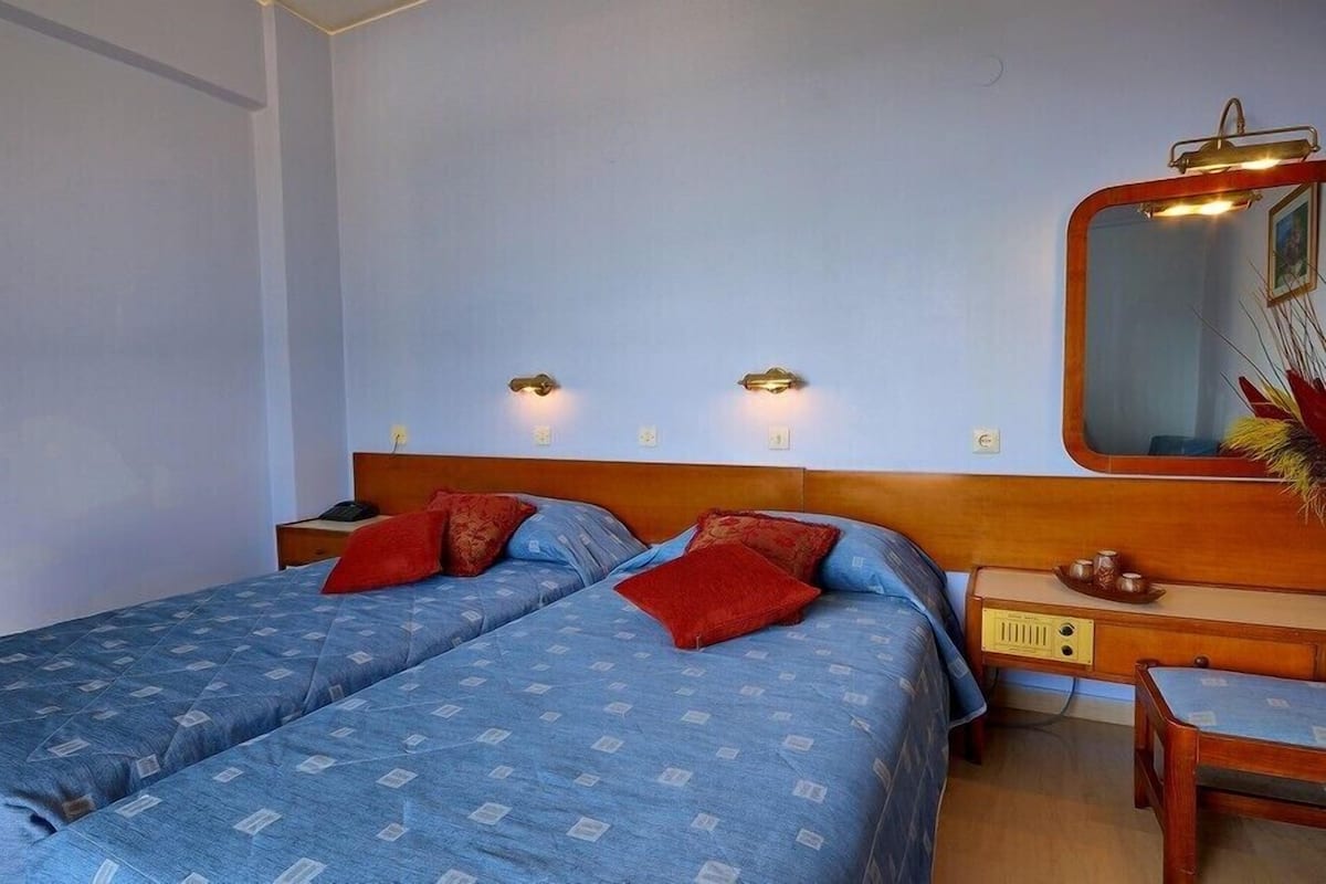 A room with two beds, a wooden headboard, bedside tables, a mirror, and wall-mounted lights.