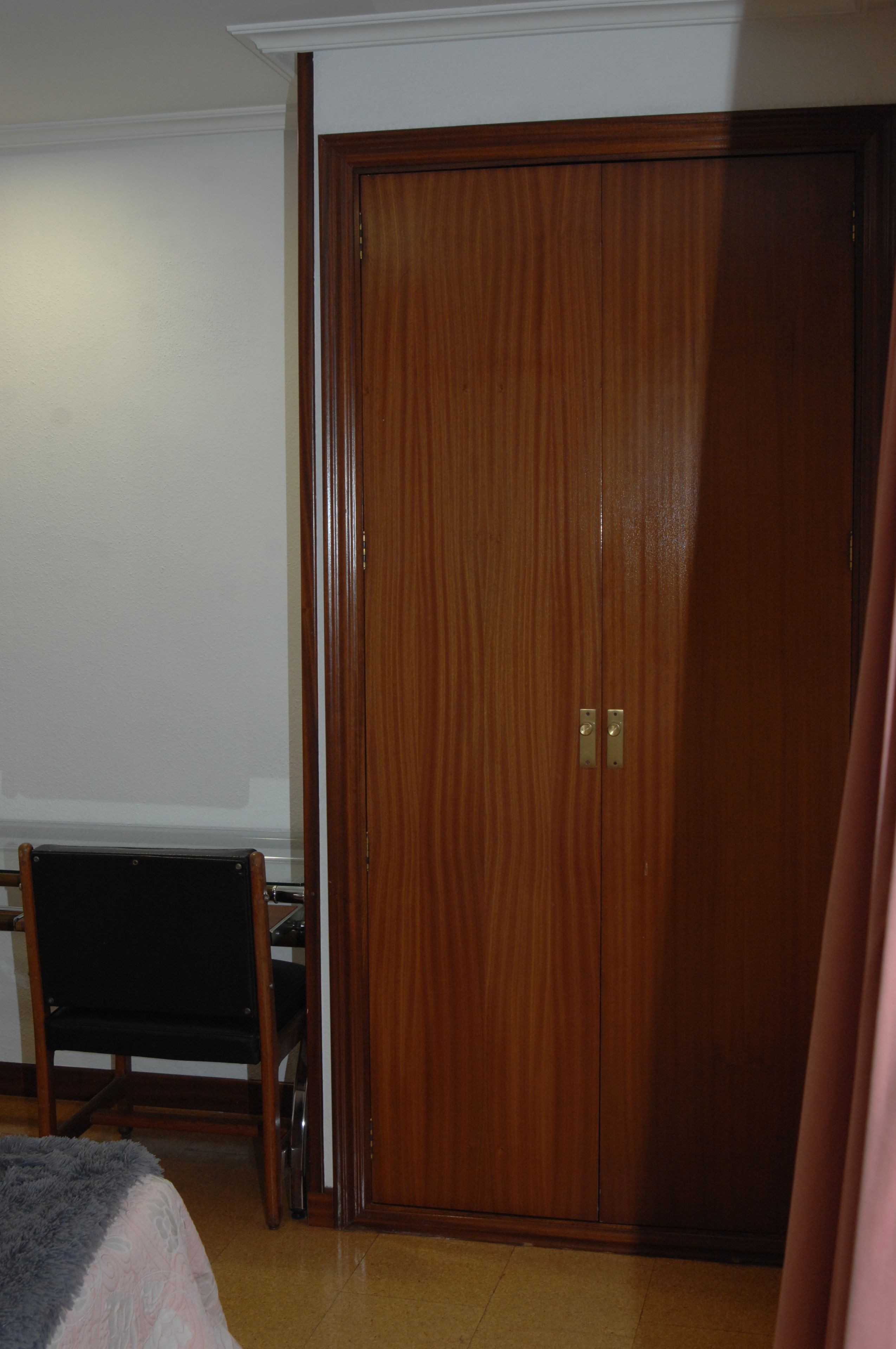 Ein Zimmer mit einem Holzschrank, einem schwarzen Stuhl und einem Bett mit grauer und pinkfarbener Decke.