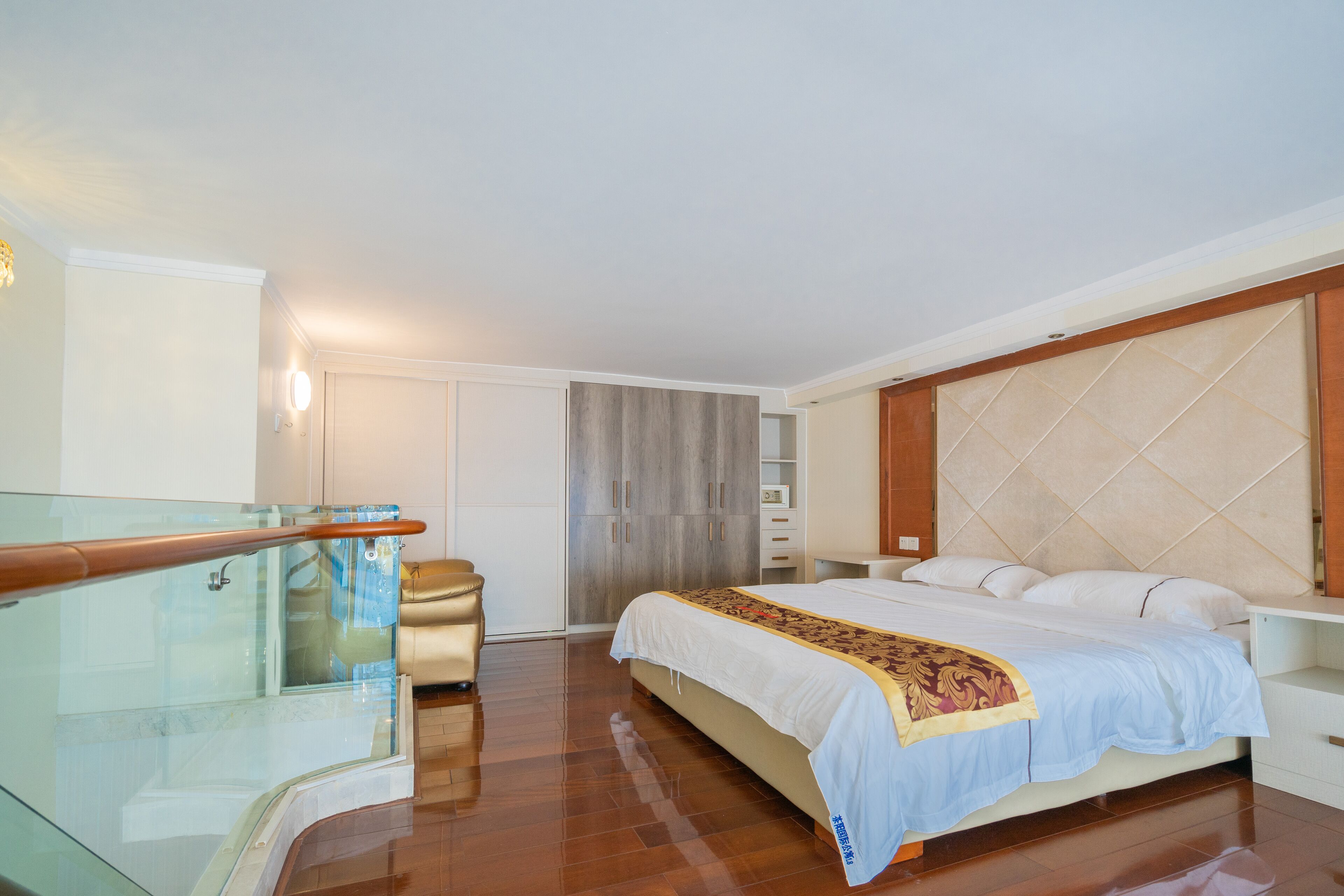 Une chambre à coucher comprenant un lit, une tête de lit en bois, une rambarde en verre, une armoire et un petit coin salon.