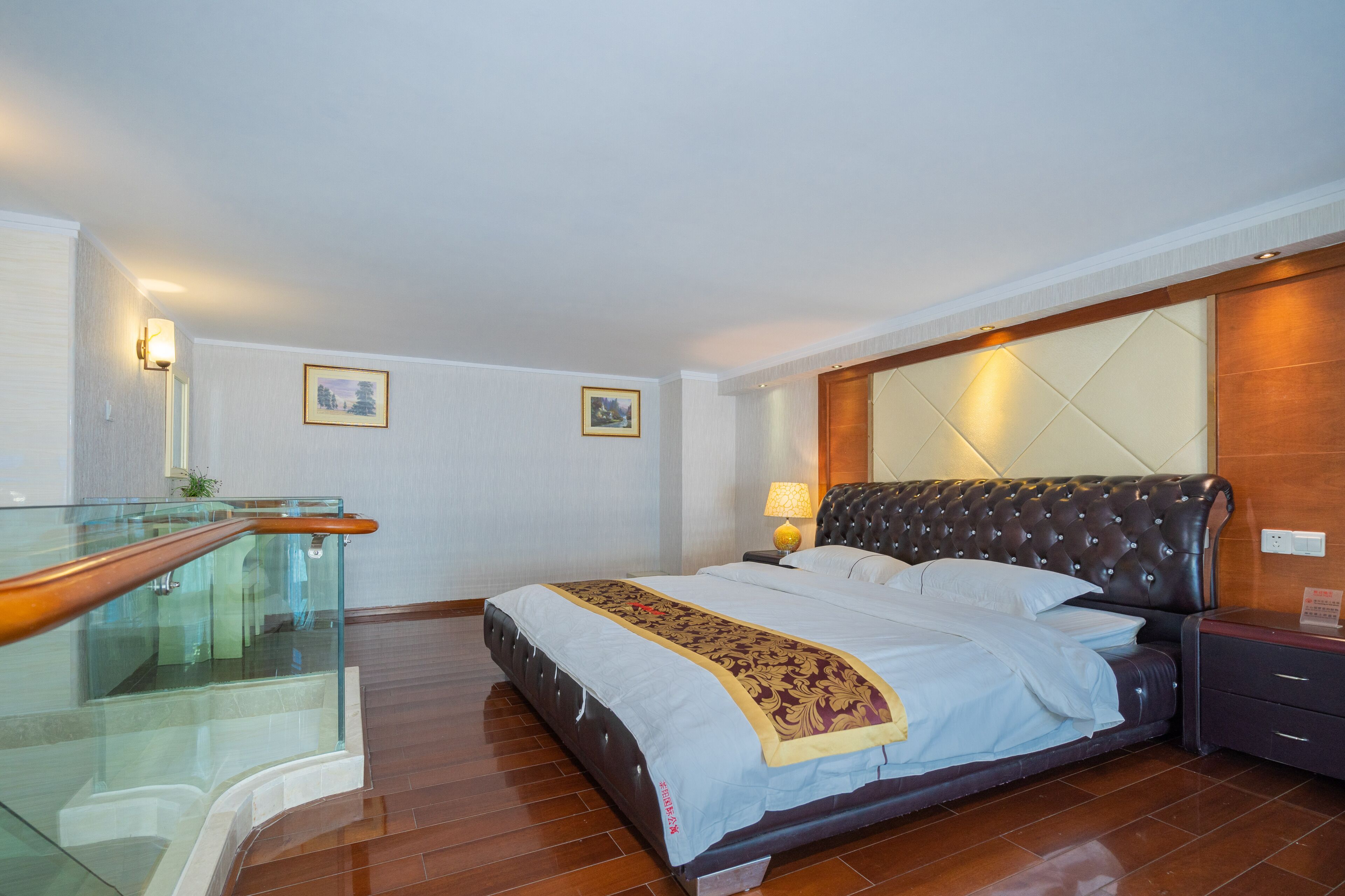 A hotel room with a large bed, a glass railing, a wooden headboard, and a nightstand with a lamp.