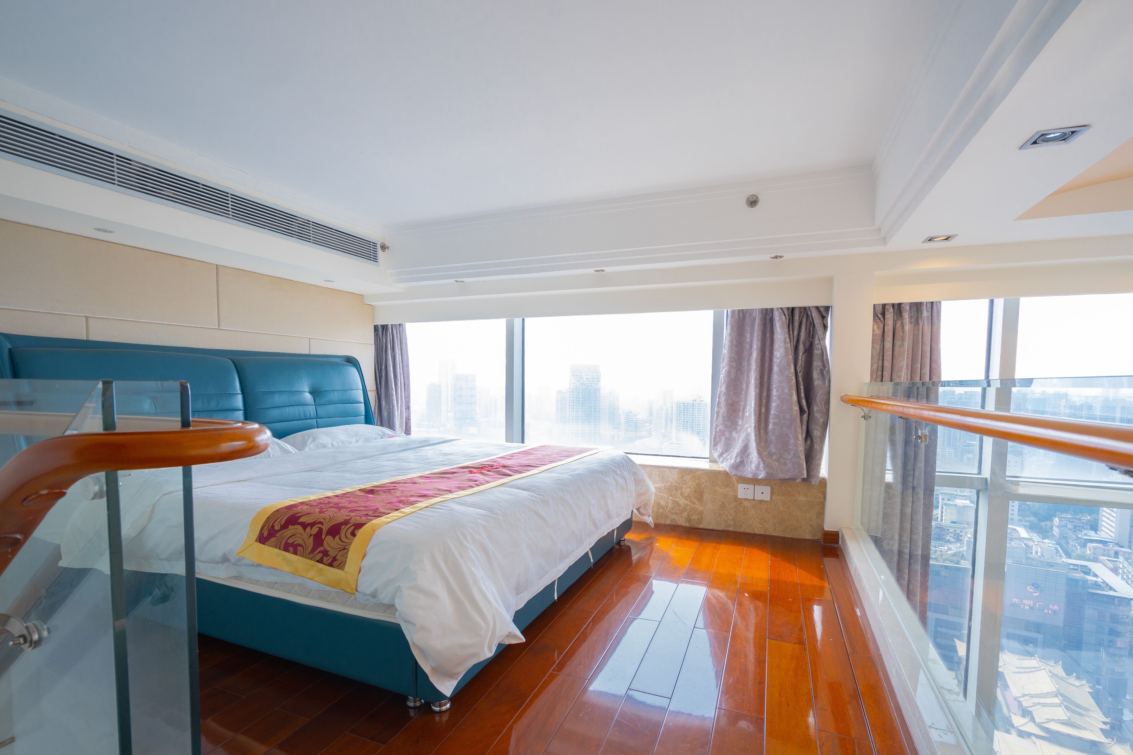 A modern hotel room with a large bed, a balcony with a glass railing, and a city view.